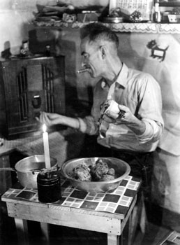 “Helping the cook.” Claude multi-tasks by peeling potatoes, tuning the radio, and smoking a cigar at the cabin in Twelve Mile. 1938